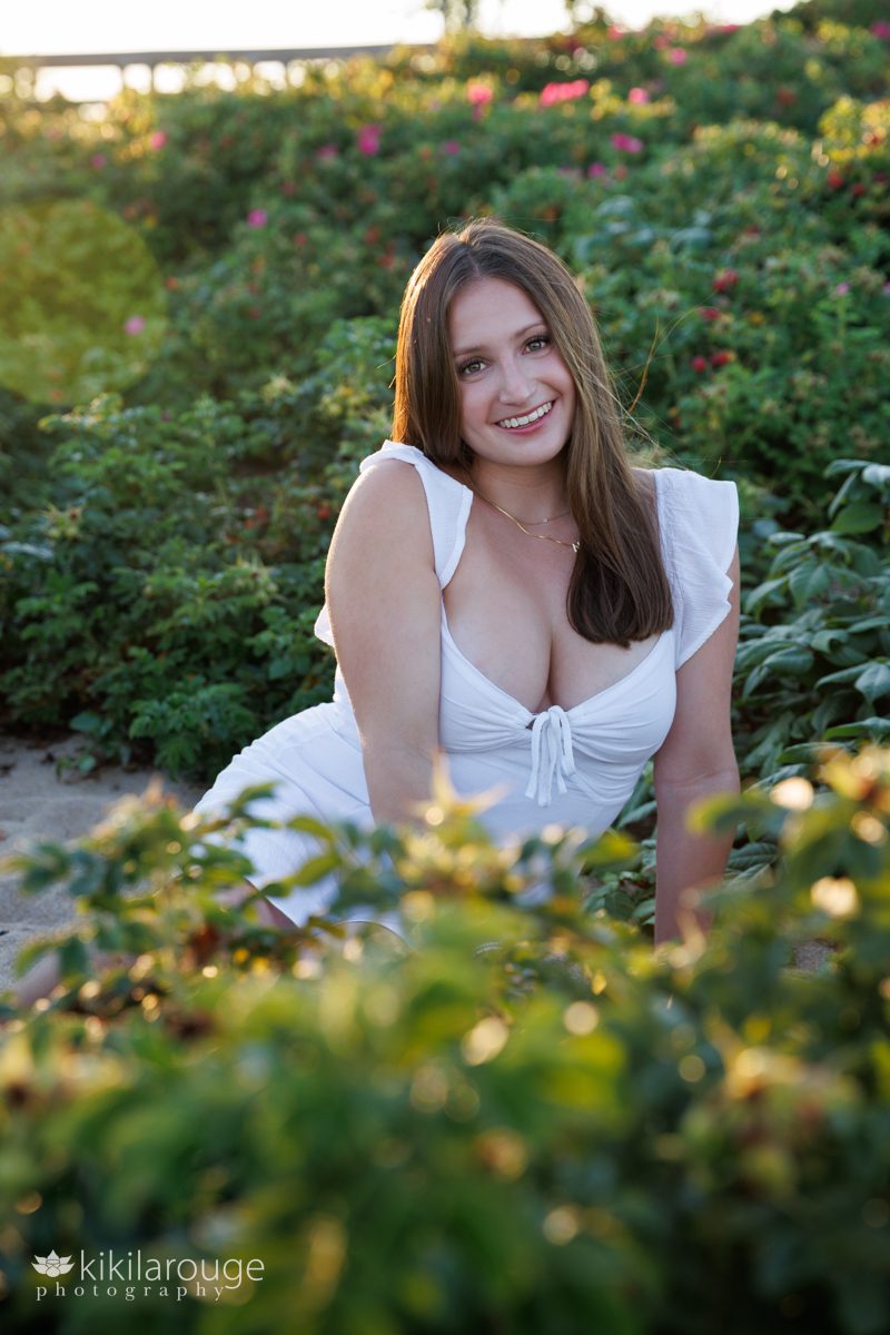 Young woman in white dress smiling with sun flare sitting on sand surrounded by beach roses