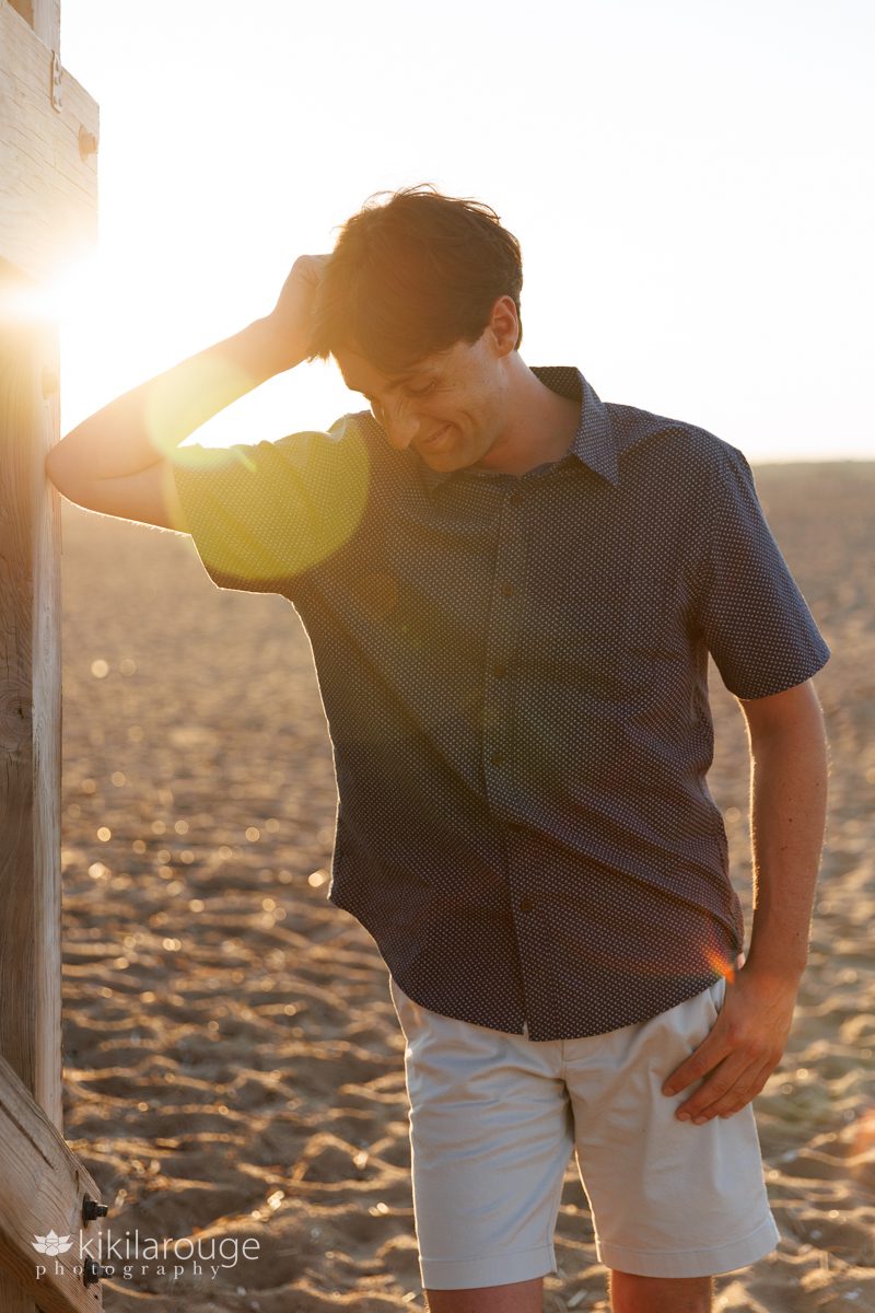 Young man in blue button up shirt looking down laughing leaning on wooden beach boardwalk with sun flare