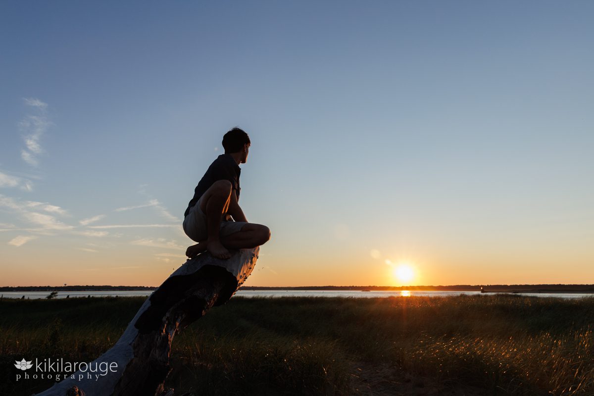 Young man sitting atop a piece of large driftwood looking out at the sunset in silhouette