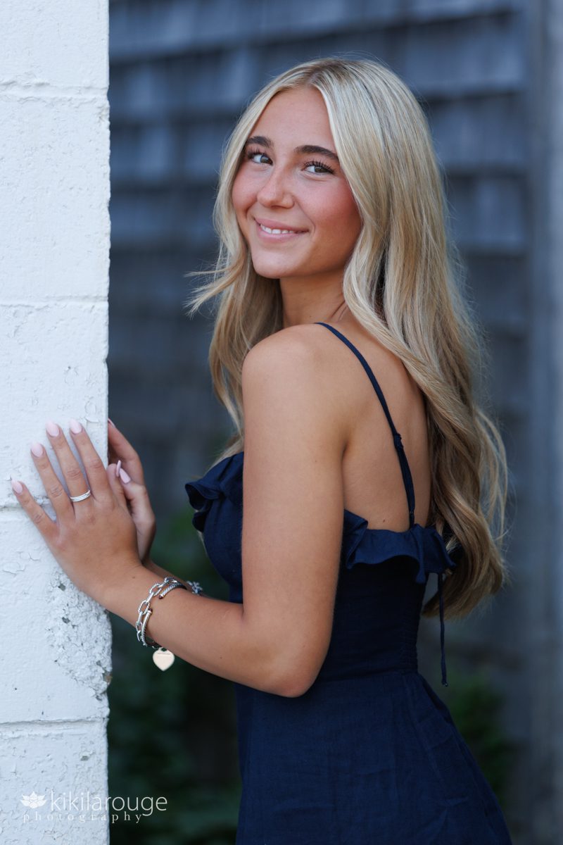 Senior girl long blonde hair in navy blue dress leaning agains a white wall smiling