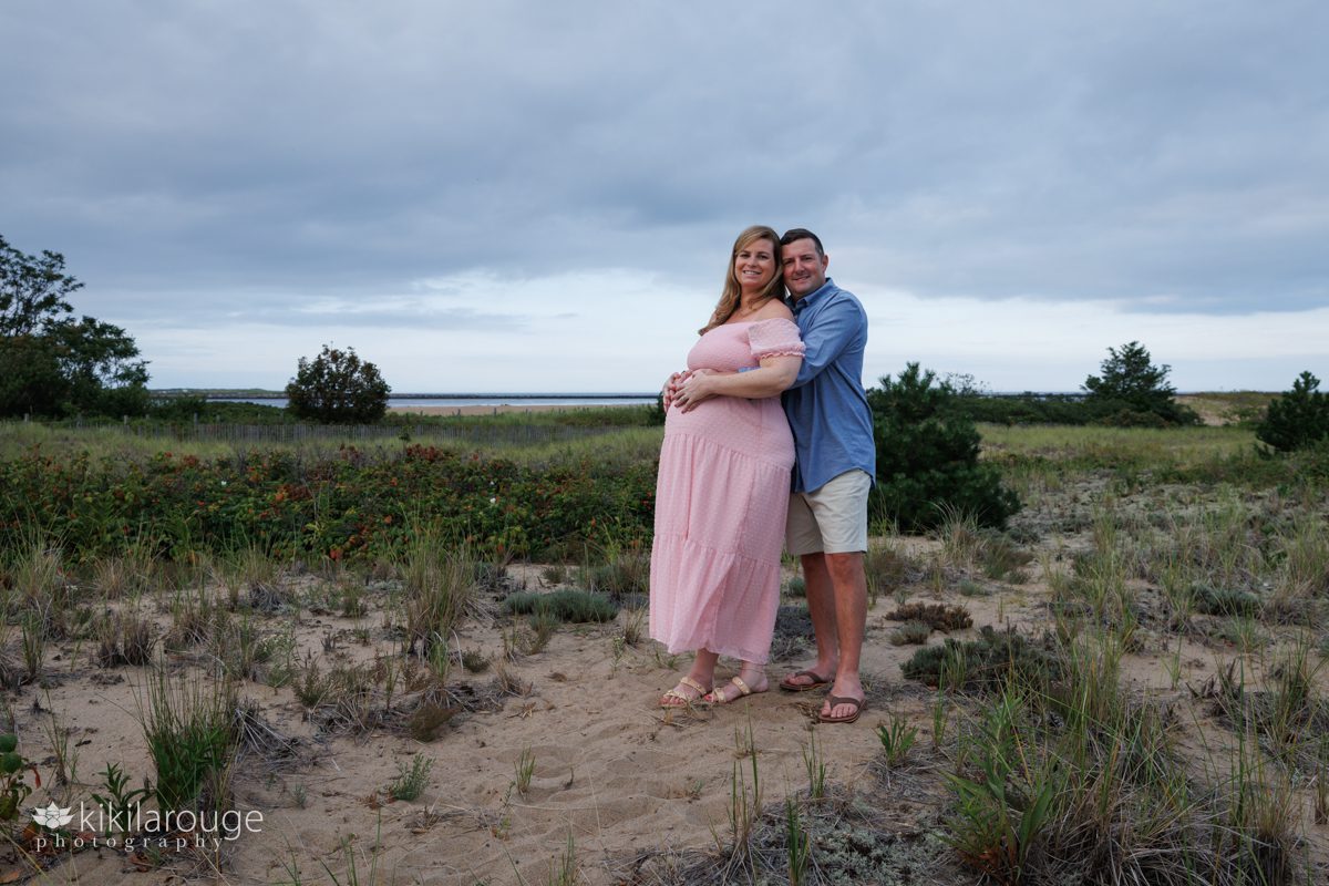 Pregnant woman in long pink dress and man in blue shirt hugging her belly in the dunes of Plum Island with beach and water in the backdrop