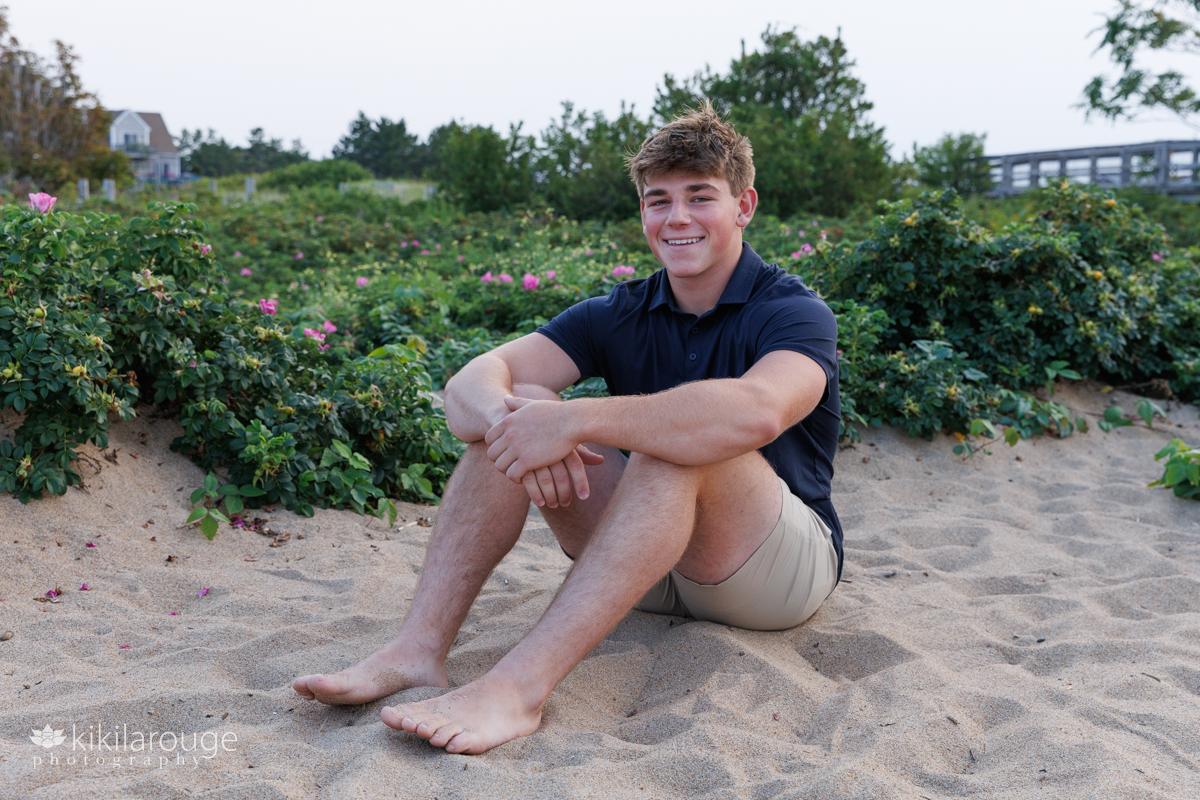 Senior boy sitting in sand with khaki shorts and navy blue shirt smiling