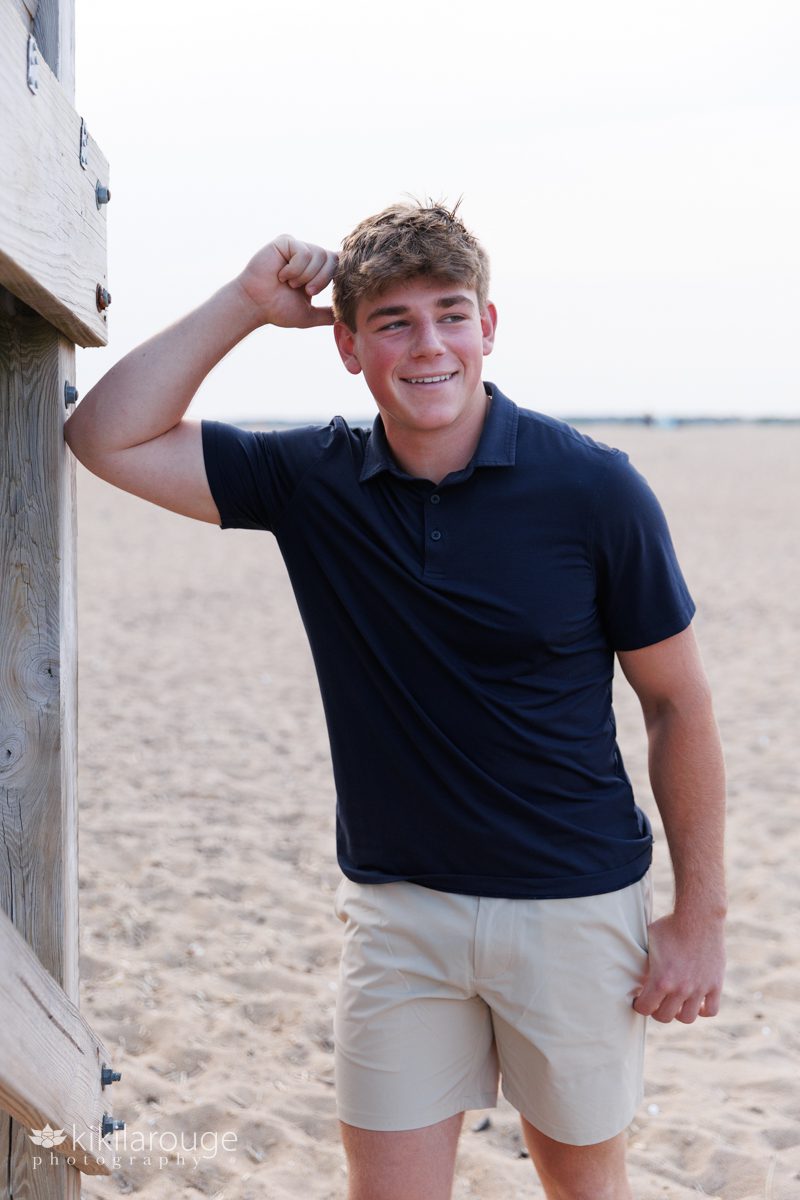 Senior boy leaning on wooden boardwalk at Plum Island beach dressed in khaki shorts and a navy polo shirt