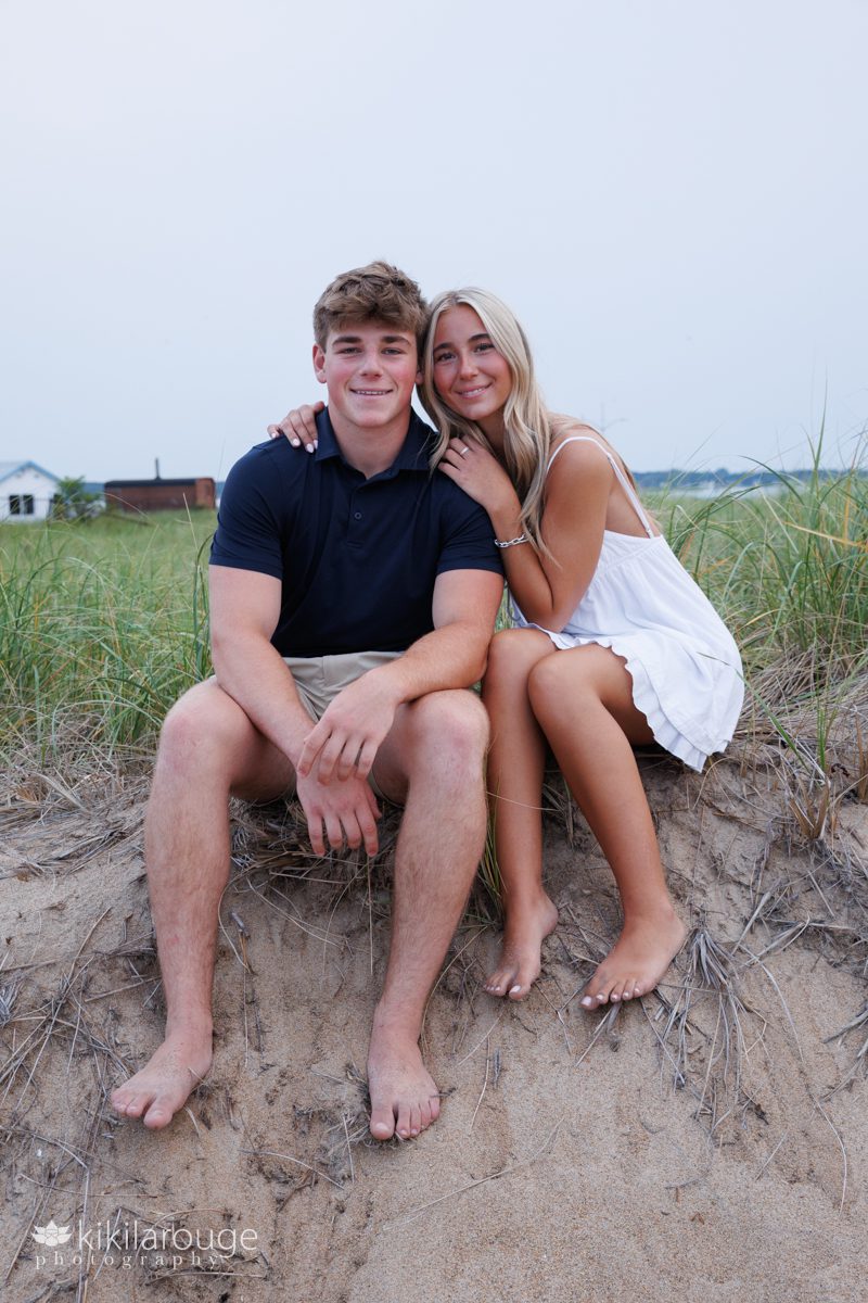 Irish twin Senior siblings a girl with long blonde hair in a white sundress and boy in khaki shorts and navy blue shirt sitting on dune edge hugging
