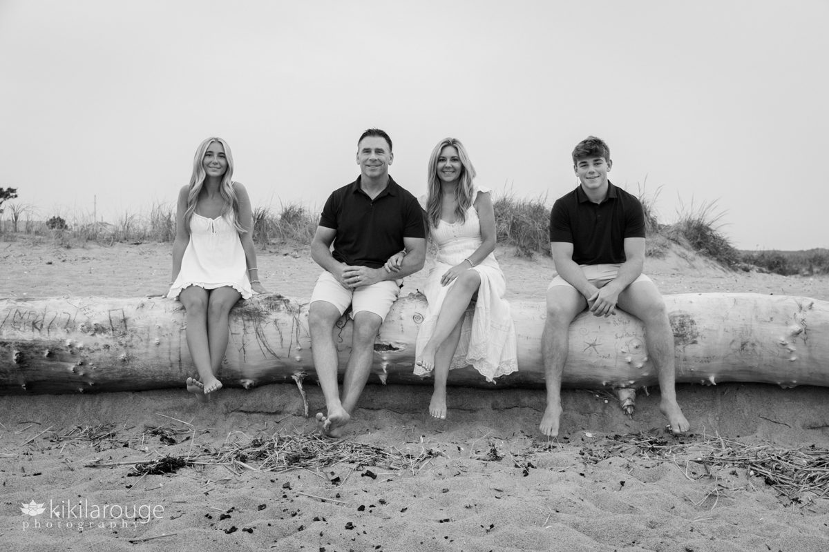 Family of four sitting on large white driftwood at Plum Island beach with Mom and Dad in the middle and Senior boy and girl on either side
