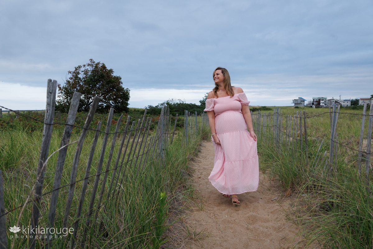 Pregnant woman walking down dune beach path in long pink dress smiling and looking off to the side