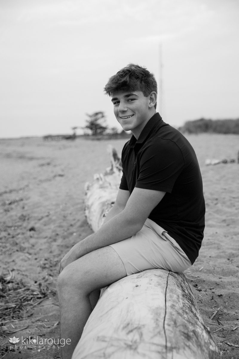 Senior boy sitting on large driftwood at beach looking and smiling into the camera
