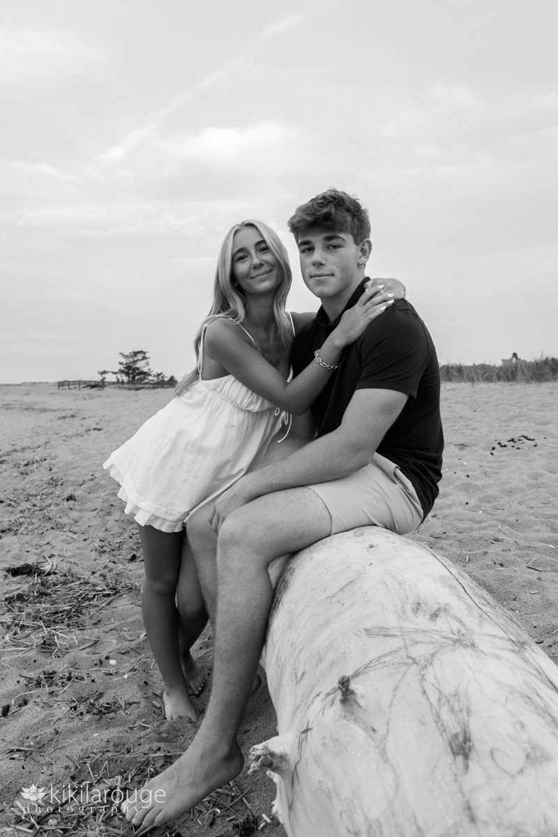 Senior siblings with girl hugging her brother who is at the beach sitting on large piece of driftwood