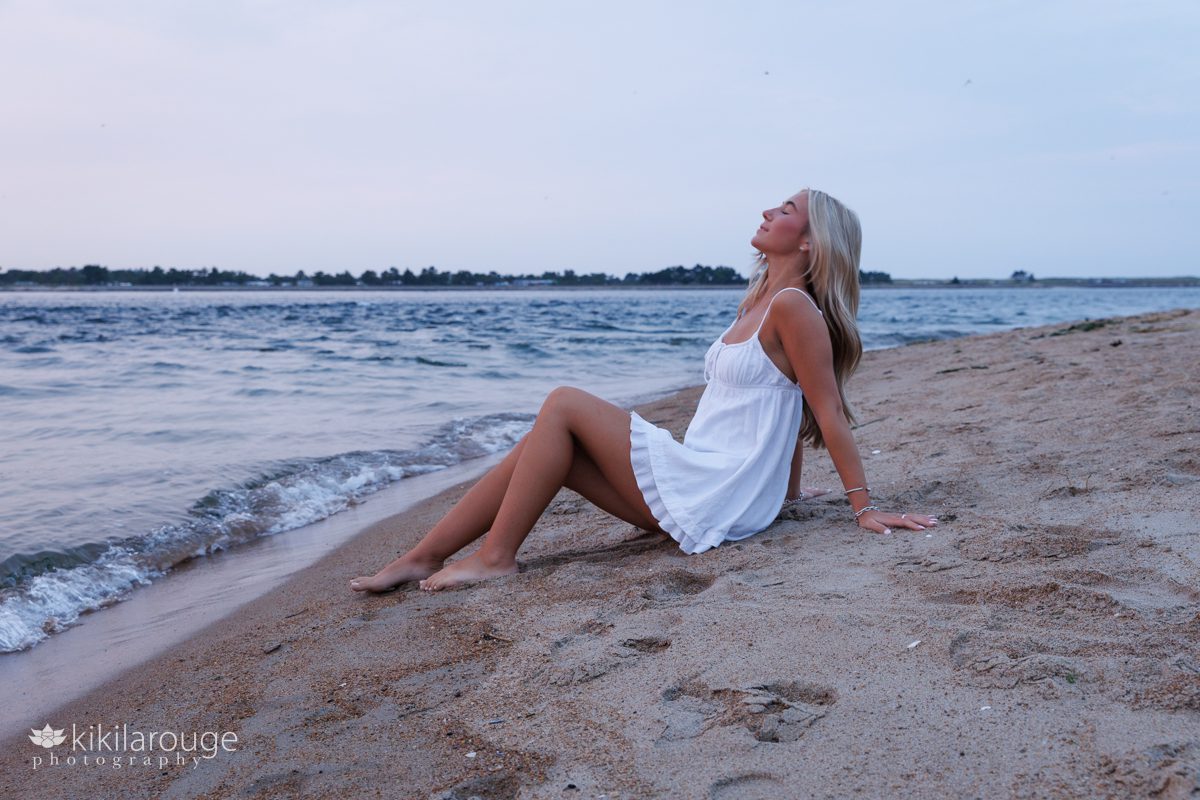 Senior girl sitting at water's edge at the beach tilting her head back with eyes closed at sunset