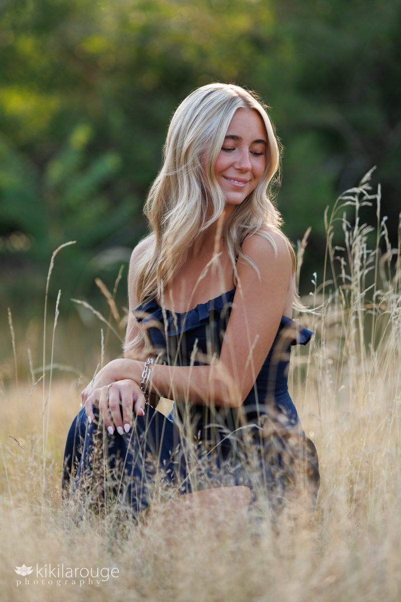 Senior girl long blonde hair in navy blue dress looking down sitting in a beach meadow