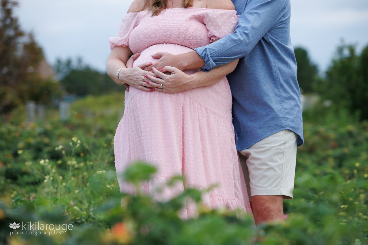 Close up image of a man and woman holding hands atop a pregnant belly woman in a long pink dress