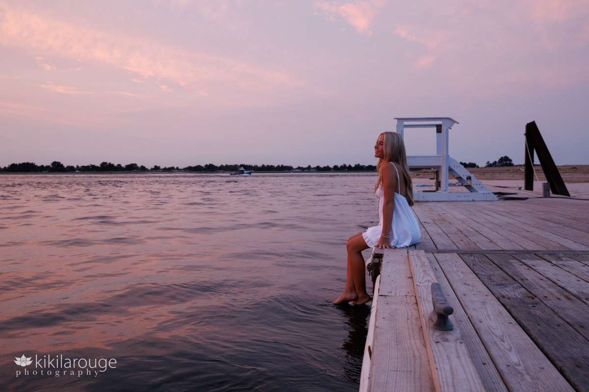 Pink sunset on the water and a girl sitting next the dock in a white sundress looking out