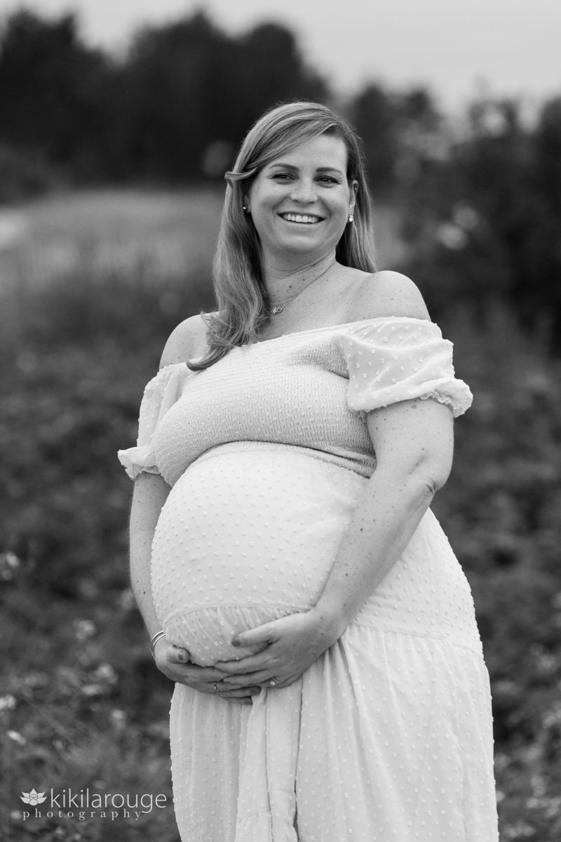 BW of pregnant women in maternity dress holding her belly laughing and looking a bit off to the right at her husband out of frame