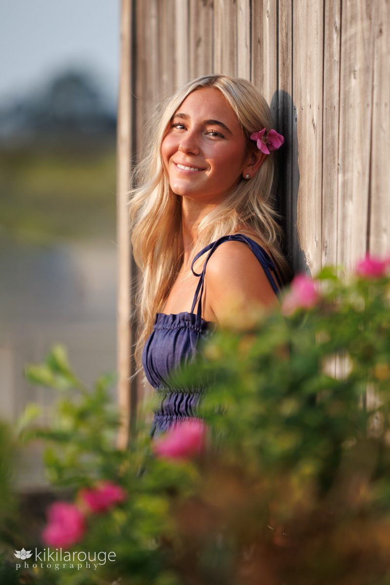 Senior girl leaning on wood wall with beach roses in the foreground and in her hair