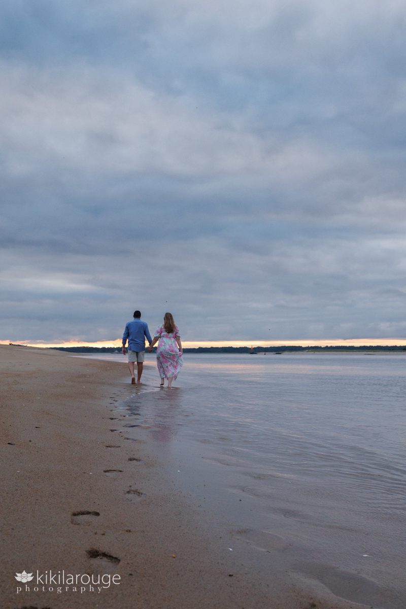 Couple holding hand walking down Plum Island beach at the shore line with dramatic skies and their footprints in the foreground