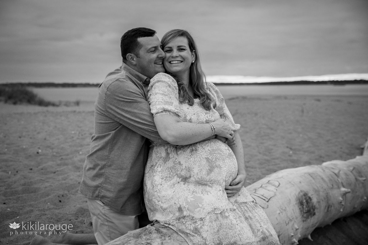 BW portrait of a man hugging his pregnant wife who is sitting on large piece of white driftwood on Plum Island beach