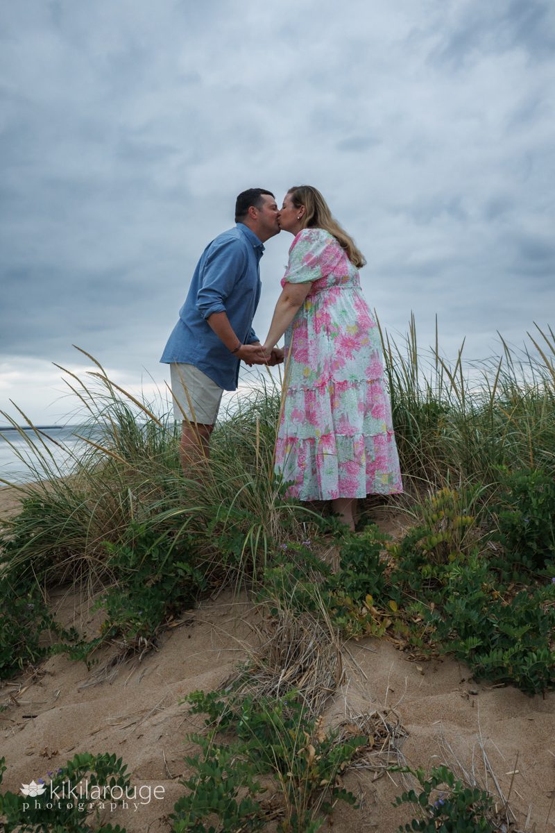 Couple standing in beach dunes holding hands facing each other leaning in for a kiss with pregnant woman in Lily Pulitzer pink and green floral dress