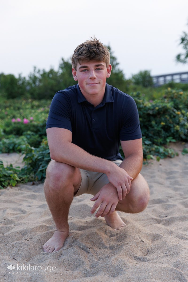 Senior boy sitting in sand with khaki shorts and navy blue shirt with a smirk