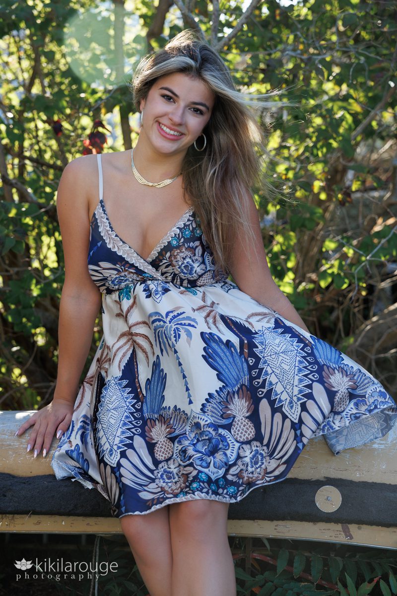 High School Senior girl in blue and brown print silk dress blowing in wind backlit by sun sitting on old canoe