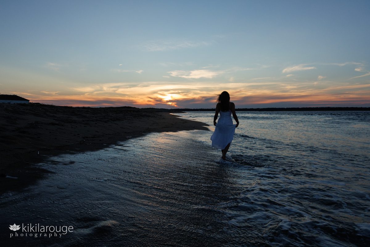 Young woman in white dress walking along the shore line with waves towards a dramatic orange and blue sunset in silhouette