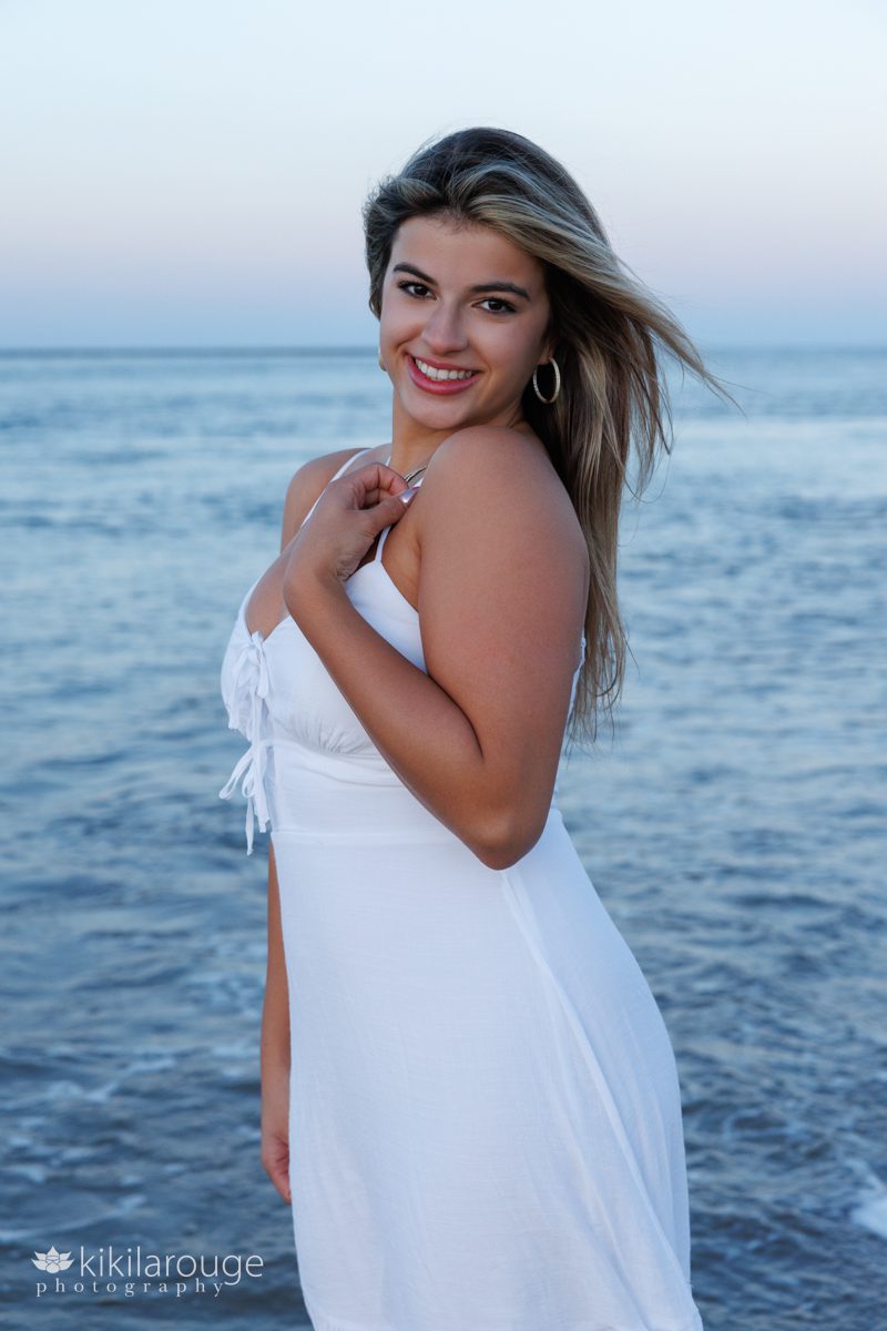 Senior picture of girl with long hair and blonde highlights in white dress with hand on decollate and blue and pink water and sky in backdrop