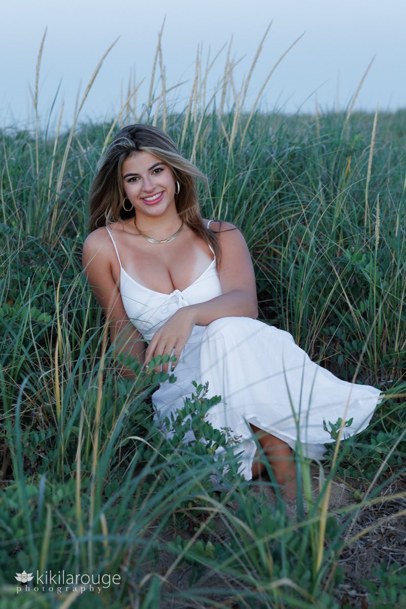 Senior pic of girl in white long summer dress sitting in beach dunes and Plum Island with wind blowing her and hair and all the dune reeds