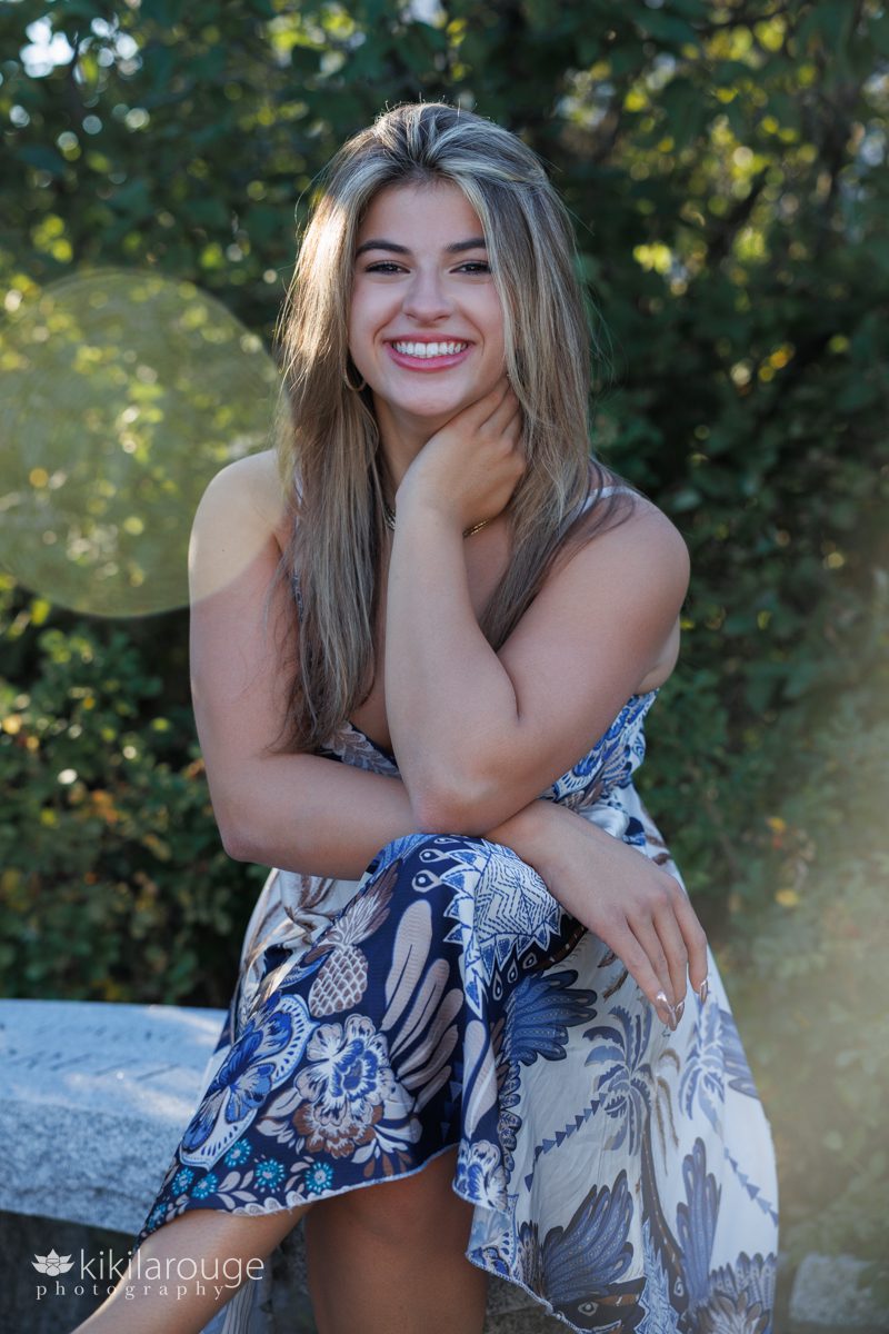 High School Senior girl in blue and brown print silk dress sitting on cement bench with hand at neck and big smile