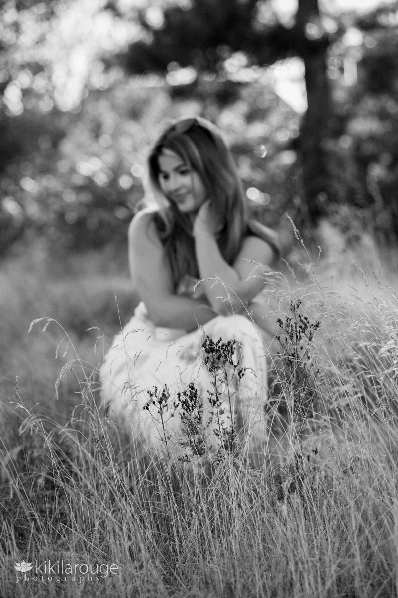 Senior girl in white dress crouched in beach reeds out of focus looking down