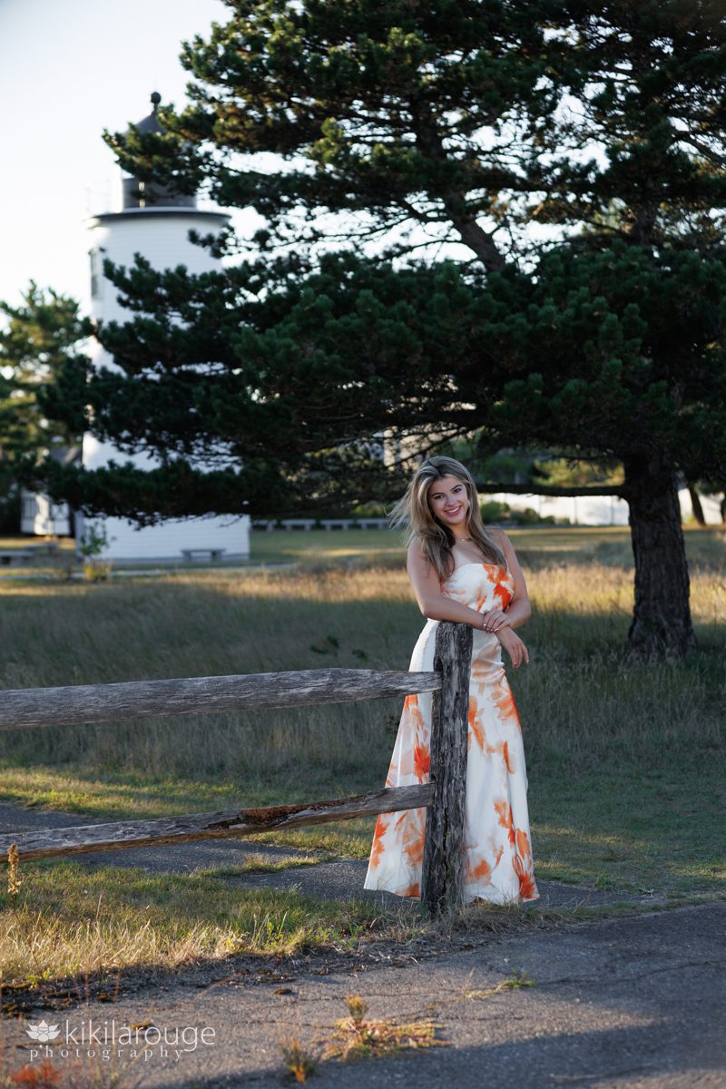 Senior girl leaning on wooden fence wearing a long white and orange dress with big tree and Plum Island lighthouse in background