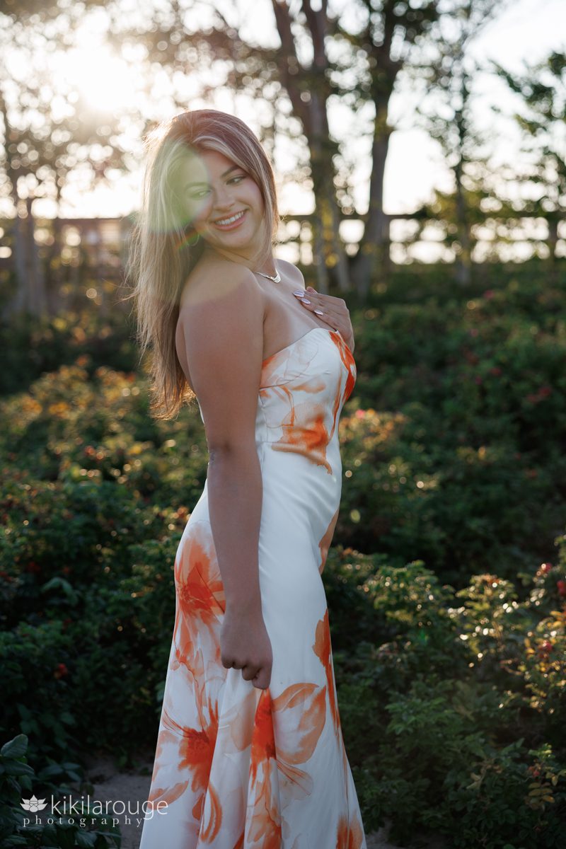 Girl standing in beach dunes with beach roses wearing long white and orange dress looking down with sun flare