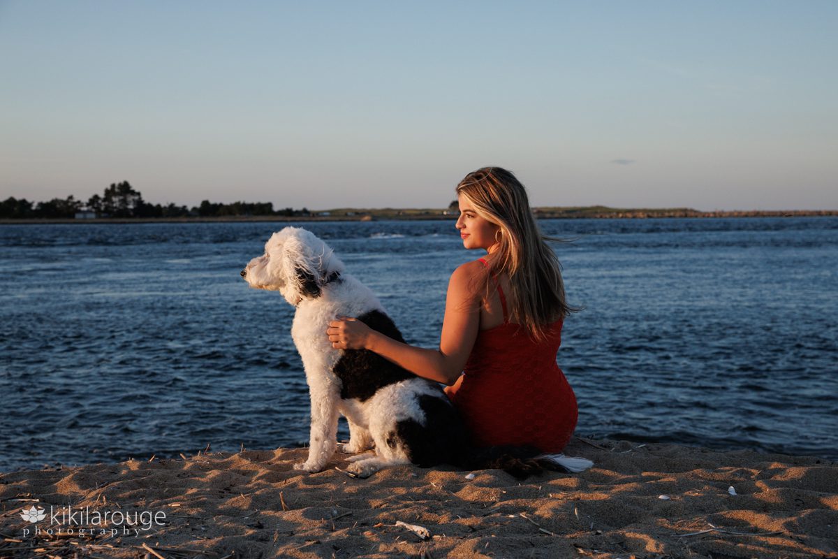 Senior girl portrait sitting at water's edge on Plum Island with her doodle dog both looking to the left into the sunset
