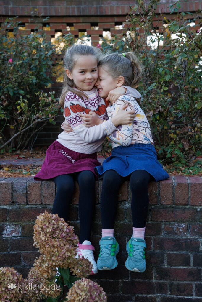 Two young sister sitting on brick wall hugging one snuggled into the other with smiling scrunched face wearing black tights and little skirts with sneakers