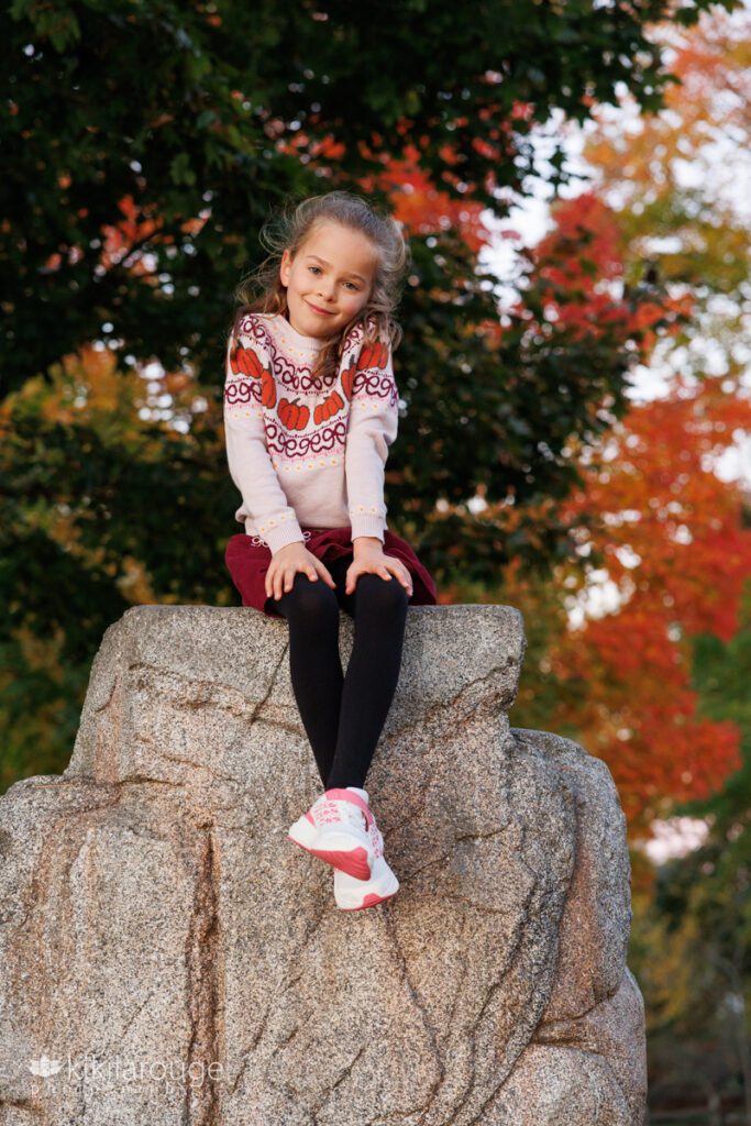 Sweet seven year old girl sitting atop a rock with legs crossed and gorgeous red and yellow foliage in backdrop