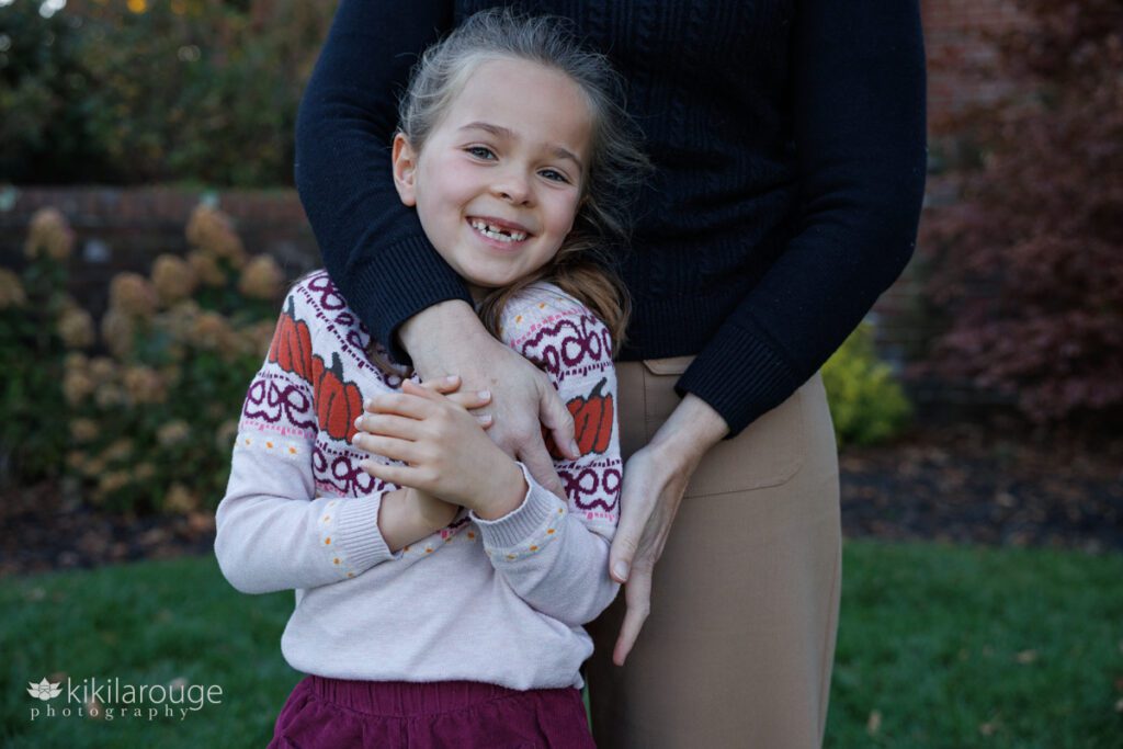 Your girl in pumpkin sweater smiling at the camera as Mom hugs her 