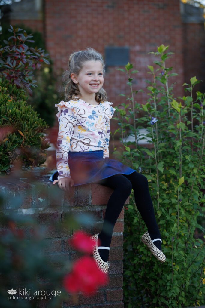 Young girl in floral shirt and denim skirt with black leggings sitting on garden brick wall looking off at Mom with flowers all around
