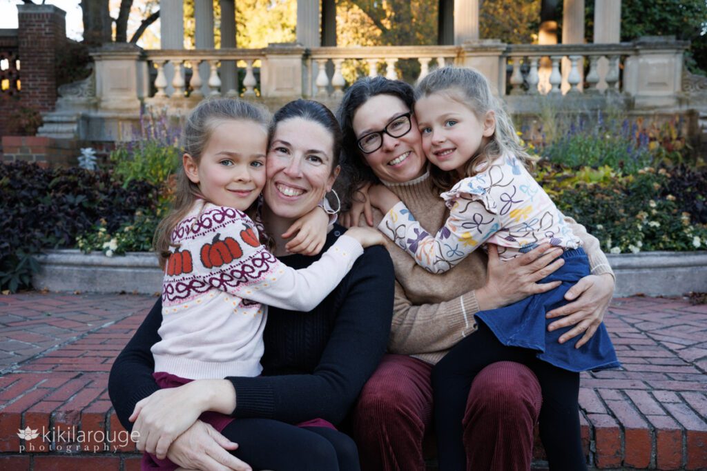 Cute family of four with two smiling Moms and their young daughters hugging them both