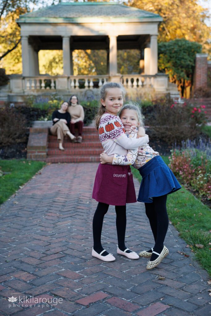 Two sisters hugging in brick rose garden with their Moms out of focus in backdrop sitting on brick steps