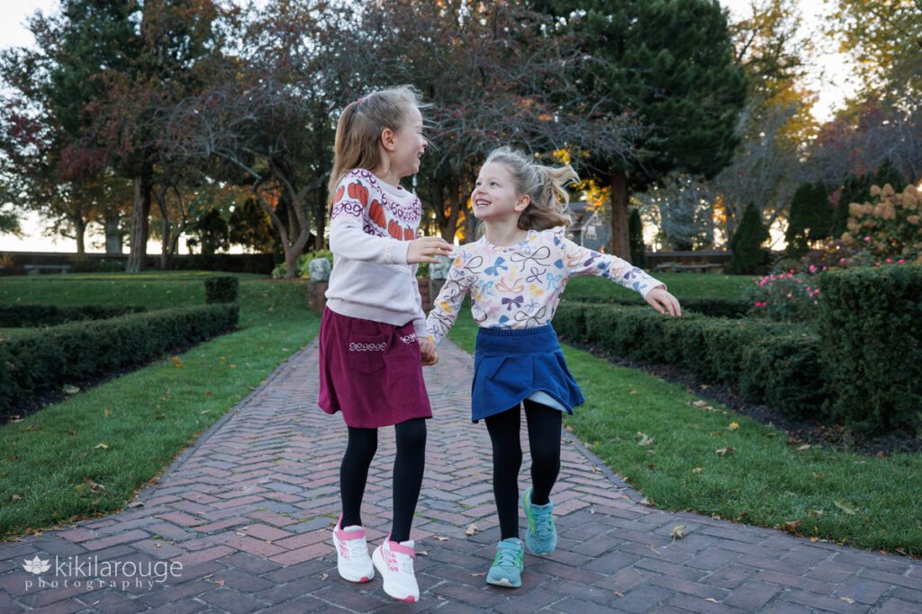 Two young girls holding hands running and looking at each other laughing and smiling in a brick rose garden at Lynch Park Beverly MA