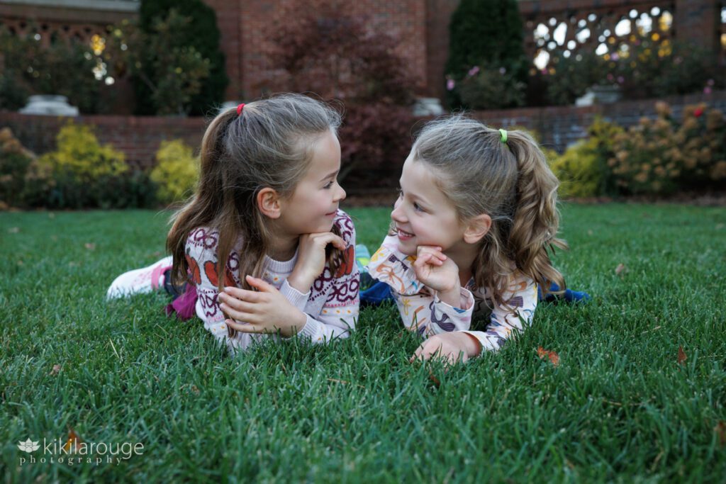 Two young sisters laying in the grass looking at each other smiling in a brick rose garden at Lynch Park Beverly 