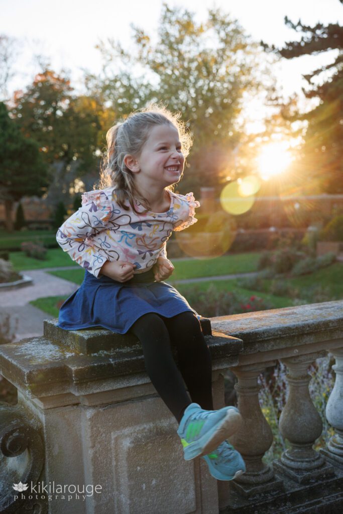 Six year old girl sitting on stone railing in Lynch Park rose garden with sun flare smiling and doubling over